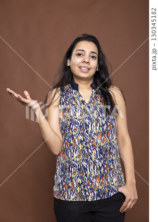 Portrait of female indian adult posing with one hand in pocket, standing against isolated background. Smiling beautiful woman in casual outfit, gesturing to side as if giving presentation in studio. 130345182