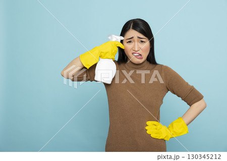 Overwhelmed asian housewife in yellow gloves holds cleaning spray bottle near forehead, expressing fatigue and frustration with household chores. Ideal for cleaning burnout concepts. 130345212