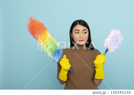Young female janitor stands prepared with colorful cleaning tools during morning routine. Confident asian housekeeper ready for household chores, posing against isolated background. 130345238