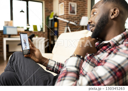 Black freelancer sitting on sofa and discussing with caucasian doctor on video call. African american man having virtual meeting on phone with professional physician, working on medical business ideas 130345263