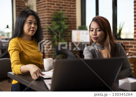Businesswoman holding a coffee cup collaborates with her colleague at desk, preparing project presentation in brick wall office. Female marketing specialists reviewing company information on laptop. Businesswoman holding a coffee cup collaborates with her colleague at desk, preparing project presentation in brick wall office. Female marketing specialists reviewing company information on laptop. 130345264