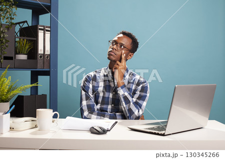 African american manager with his hand on his cheek, looking above and having deep thoughts, reflecting on office reports. Black man sitting at desk, touching his face and thinking of business ideas. 130345266
