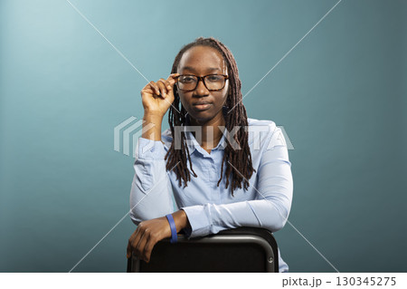 Young african american woman touching her glasses, posing confidently isolated over blue background. Black female model sitting on chair and looking at camera with focused expression. 130345275