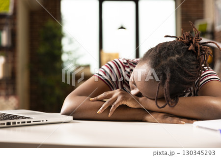 African American female entrepreneur sleeping at desk after overworking on laptop. Tired black businesswoman resting head on her arms, taking nap to recover from long hours at home. African American female entrepreneur sleeping at desk after overworking on laptop. Tired black businesswoman resting head on her arms, taking nap to recover from long hours at home. 130345293