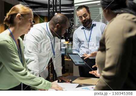 Multiethnic project team shares analytics reports during advisory board briefing late at night, reviewing business intel and preparing metrics for upcoming presentation with shareholders. 130345308