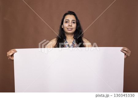 Portrait of indian woman holding empty banner placard, standing in front of brown background. Beautiful female individual posing with blank white poster, ideal for showcasing promotional material. 130345330