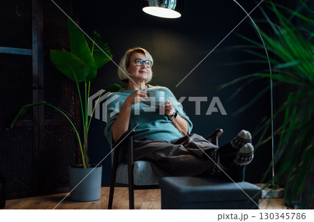Middle aged woman relaxing with cup of hot drink in scandy style hygge interior home. Cozy comfortable winter or autumn weekends. Take a break and relax. Lady enjoy calm mood, well being alone. 130345786