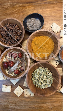 Colorful spices like turmeric, star anise, cardamom, and cinnamon sticks arranged in wooden bowls on a wooden surface create a vibrant culinary scene with scattered crackers 130345880