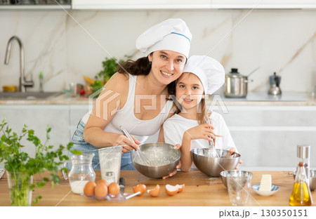 Mom and girl in white chief hats are talking in kitchen and whisking ingredients for pastry in bowl 130350151