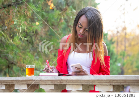 Woman in a red jacket writing notes while enjoying tea in a park during autumn 130351893