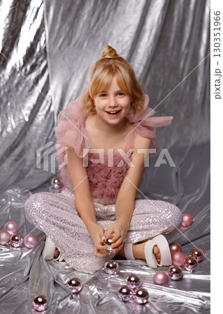 Smiling blonde girl in pink dress sitting with Christmas balls on silver background 130351966