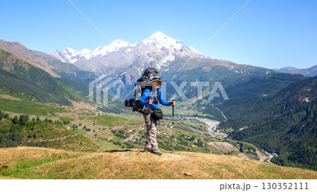 Hiker stands proudly on a mountain summit with snow-capped peaks in the background 130352111