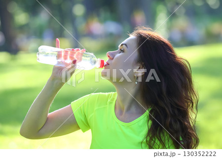 Woman enjoying hydration in a park during sunny weather while wearing athletic clothing 130352222