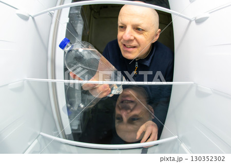 Man examines bottle of water in open refrigerator during mid-morning 130352302