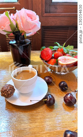 Porcelain coffee cup with creamy milk, chocolate bonbon resting on white saucer, wooden surface accented by soft pink roses, fresh berries in muted background Porcelain coffee cup with creamy milk, chocolate bonbon resting on white saucer, wooden surface accented by soft pink roses, fresh berries in muted background 130354630