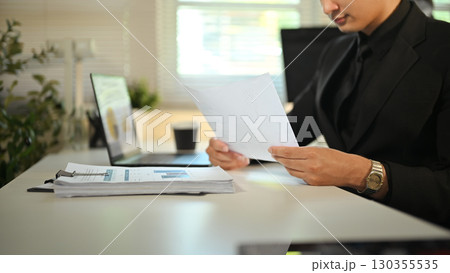 Businessman in formal suit reviewing financial documents at modern office desk Businessman in formal suit reviewing financial documents at modern office desk 130355535