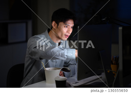 Busy businessman working late at night, eating instant noodles at desk while looking at computer screen 130355650