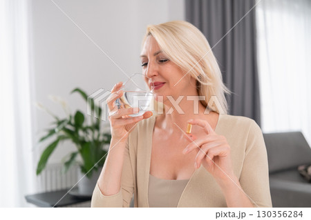 Blonde woman holding pill in one hand and glass of water in the other in modern living room, daily health practice  130356284
