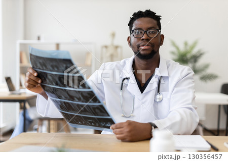 Male African doctor wearing white coat with stethoscope analyzing X-ray in modern office during daytime, demonstrating care for patient health and diagnosis processing Male African doctor wearing white coat with stethoscope analyzing X-ray in modern office during daytime, demonstrating care for patient health and diagnosis processing 130356457