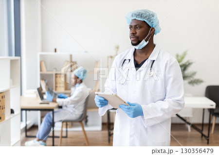 Male doctor in medical uniform using tablet in modern office environment alongside female coworker. Male doctor in medical uniform using tablet in modern office environment alongside female coworker. 130356479