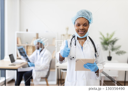 Young African female doctor wearing uniform and mask, holding tablet, giving thumbs up gesture. Smiling expression conveys confidence and professionalism. Background colleague working on patient files Young African female doctor wearing uniform and mask, holding tablet, giving thumbs up gesture. Smiling expression conveys confidence and professionalism. Background colleague working on patient files 130356490
