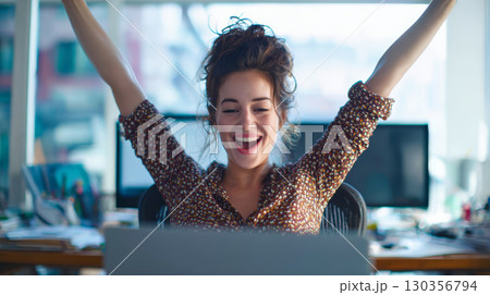 Young woman celebrating success with raised arms in office, smiling happily while sitting at desk with computer Young woman celebrating success with raised arms in office, smiling happily while sitting at desk with computer 130356794