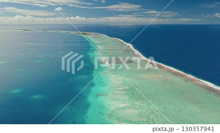 Crystal clear turquoise waters stretch out around a narrow sandy islet under a sunny sky. The coastline features stunning coral reefs that teem with aquatic life. 130357941