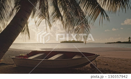 Small boat resting on sandy shores of a tropical beach at sunset, nestled under the shade of a palm tree, while a picturesque island emerges in the distance 130357942