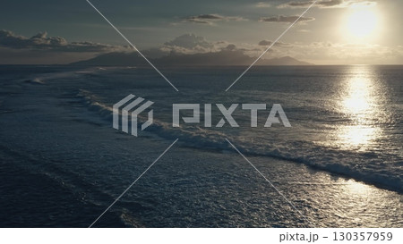 Golden sunset light reflects on the surface of a calm ocean as gentle waves break near a tropical island, creating a serene and idyllic scene 130357959