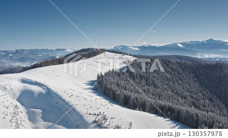 Breathtaking aerial view of a snow covered mountain range with dense coniferous forest, serene and picturesque winter landscape under a clear blue sunny sky. Winter wild nature travel background Breathtaking aerial view of a snow covered mountain range with dense coniferous forest, serene and picturesque winter landscape under a clear blue sunny sky. Winter wild nature travel background 130357978