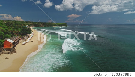 Stunning aerial drone highlights Dreamland Beach in Bali, Indonesia, with turquoise waves gently crashing on the sandy shore, vibrant beach umbrellas, and a red beach hut 130357999