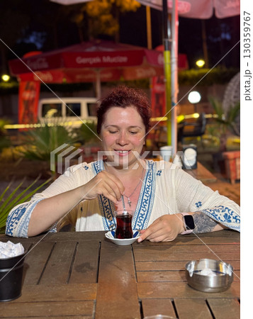 Woman stirring Turkish tea in a traditional glass at a cafe table. Evening leisure, cultural travel, and authentic lifestyle during vacation. 130359767