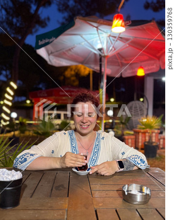 Woman sitting at an outdoor cafe table with a glass of Turkish tea. Leisure, travel, and authentic cultural experience during evening relaxation. 130359768