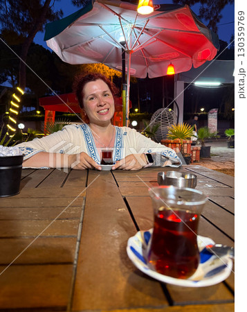 Woman sitting at an outdoor cafe table with a glass of Turkish tea. Leisure, travel, and authentic cultural experience during evening relaxation. 130359769
