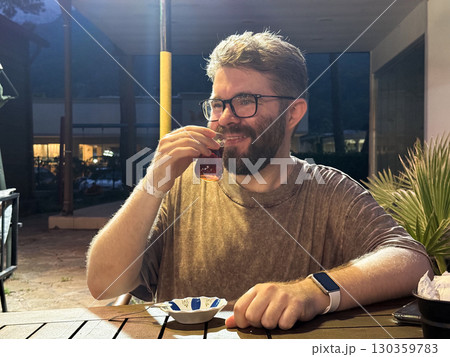 A smiling man drinks tea from traditional glass while sitting at evening cafe. Leisure, culture and everyday lifestyle in warm social environment. 130359783