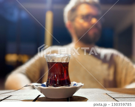 A glass of Turkish tea stands on wooden table in focus with man blurred behind. Culture, lifestyle and daily tradition of warm drink in social evening setting. 130359785