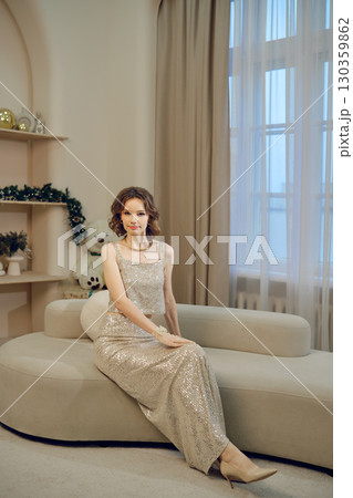 Elegant woman in a sparkling dress seated on a stylish sofa in a cozy indoor setting 130359862