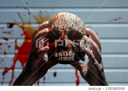 Hand holding a human skull head with blood dripping down its surface. The human skull, devil, evil, or zombie head. Scene of blood splattered on the wall. Halloween concept Hand holding a human skull head with blood dripping down its surface. The human skull, devil, evil, or zombie head. Scene of blood splattered on the wall. Halloween concept 130360099