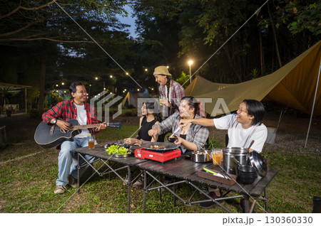Indonesian southeast asian people cooking sliced marinated beef bulgogi on a grill pan. One of them is holding an acoustic guitar. Scene of the lifestyle of enjoying food in a natural setting 130360330