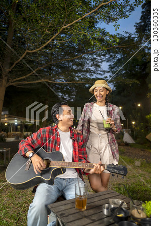 Indonesian southeast asian man playing an acoustic guitar outdoors with his friends. Showing a relaxed and enjoyable lifestyle. The scene of the lifestyle of enjoying camping in a natural setting 130360335