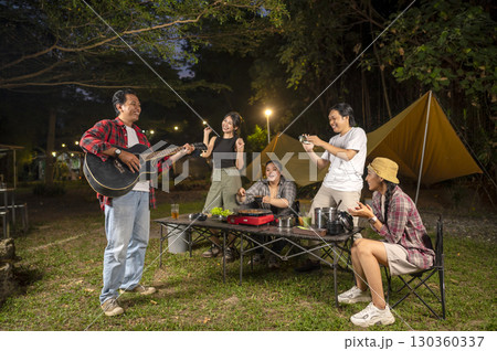 Indonesian southeast asian people cooking sliced marinated beef bulgogi on a grill pan. One of them is holding an acoustic guitar. Scene of the lifestyle of enjoying food in a natural setting 130360337