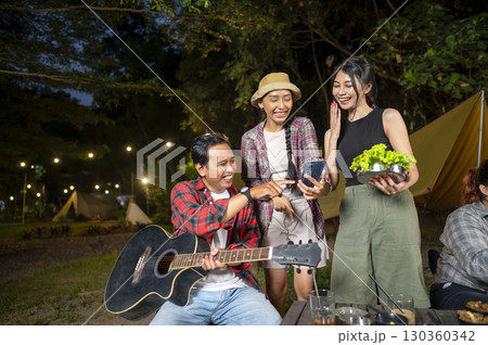 Indonesian southeast asian people enjoying quality time together. Man holds a guitar, and the other women show something on her cellphone. Scene of a lifestyle of enjoying camping in a natural setting Indonesian southeast asian people enjoying quality time together. Man holds a guitar, and the other women show something on her cellphone. Scene of a lifestyle of enjoying camping in a natural setting 130360342