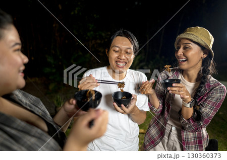 Indonesian southeast asian people are enjoying a meal, using chopsticks to serve themselves a barbecue meal from small black bowls. The scene of the lifestyle of enjoying food in a natural setting 130360373