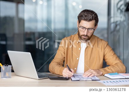 A focused businessman in glasses reviews documents at his desk, highlighting a concept of financial analysis. 130361278