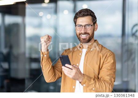 A happy man with a beard looks at his phone with a fist raised in victory. 130361348