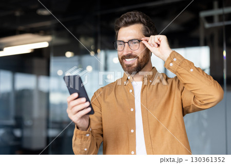 Smiling man wearing glasses and casual attire uses a smartphone in a bright, modern office space, showcasing connectivity, confidence, and effective communication. 130361352