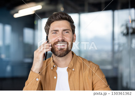 A smiling man with a beard is on a phone call, indoors with a blurred background. 130361362
