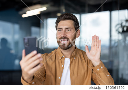A man waves during a video call on his smartphone inside an office setting, smiling and looking at the screen. 130361392