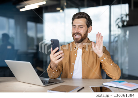 Bearded professional waving during a video call working on a smartphone and laptop in a bright, modern office. Interaction represents remote communication, technology, and professional networking. 130361394