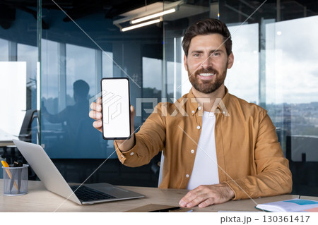 A smiling man in a brown shirt holds up a smartphone with a blank white screen, suitable for displaying an app, surrounded by office elements. A smiling man in a brown shirt holds up a smartphone with a blank white screen, suitable for displaying an app, surrounded by office elements. 130361417